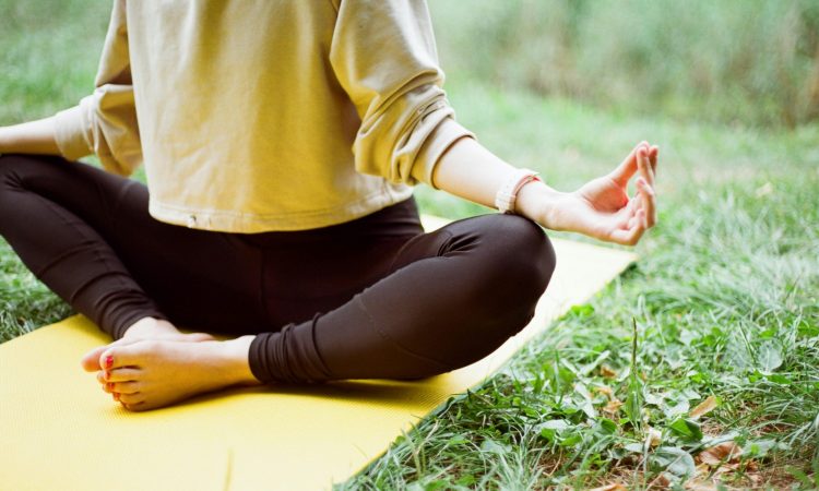 Halvkropp av kvinna sittandes i meditation ute i gräset på en gul yogamatta